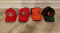 Photo showing four brand new college hats arranged side by side on a wooden surface. The hats have different colors and university logos: UNLV, University of Georgia, University of Miami (two styles).