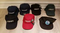 Photo showing seven brand new golf hats on a floor against wall baseboard, various colors and logos visible