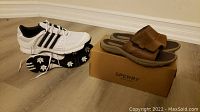 Side view of white Adidas golf shoes showing the cleated outsoles and brown Sperry sandals placed on their box.