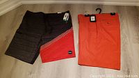 Two pairs of men's shorts side by side on floor: left is O'Neill bathing suit black with red diagonal section, right is solid orange Greg Norman golf shorts hanging on black hanger.