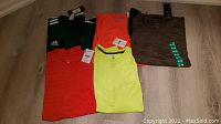 Photo showing all 5 brand new men's XL athletic T-shirts laid out on the floor with tags visible. Colors: dark green Adidas, neon yellow Nike, neon orange, solid orange and brown.