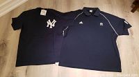 Photo of two navy blue New York Yankees shirts, a t-shirt and a golf shirt, both brand new with tags laid flat on floor.