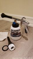 Pneumatic cleaning tool with white container, black brush attachment, rubber ring, and instructions placed on floor.