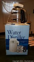 Water distiller placed on original box packaging showing the device with glass jug and white top lid, black handle visible