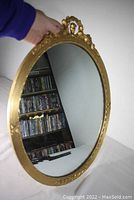 Overall view of oval mirror showing gold frame and reflection, held at an angle to display full shape and size.