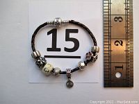 Photo showing braided leather bracelet with seven sterling silver charms and a ruler indicating bracelet length about 7 inches