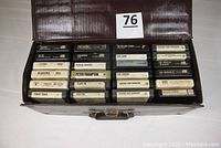 Open brown vinyl carrying case showing 8-track tapes arranged in four rows. Visible titles include AC/DC, Alabama, Bad Company, Genesis, George Benson, Peter Frampton, The Rolling Stones, The Cars, The Doors, America, Cheap Trick, Boston.