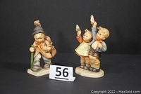 Photo of two vintage Hummel porcelain figurines against a black background showing a boy with a cello and a boy and girl waving goodbye with a lot number card 56 between them.