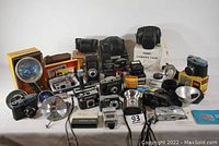 Wide view of all cameras, flashes, and cases displayed on table showing the quantity and variety in the lot including Kodak traveler folding camera, boxed Kodak Instamatic 10, Brownie StarFlash camera, multiple flashes, and cases.