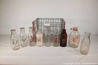 Front view of nine vintage glass milk bottles displayed in front of metal milk carrier showing variety of dairy logos and bottle styles.