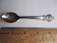 Full view of the silver-plated spoon with the spoon next to a wooden ruler showing its length is about 4.25 inches. Spoon has the word 'LUGANO' on the handle and the Rolex crown logo with 'ROLEX' at the top end.