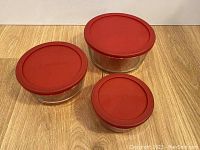 Three round glass food storage containers with red lids shown from a top angle on a wood surface, highlighting the lid shape and color.