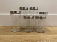 Front view of six clear glass canisters with stainless steel lids arranged on wooden surface.