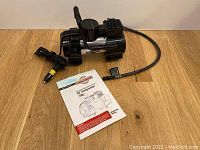 Motomaster air compressor with power cord and car lighter adapter plug, placed on wood surface alongside user manual