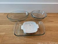 Clear glass rectangular baking dish with handles, medium oval dish, round dish with lid, and smaller white square dish with lid grouped on wooden surface.