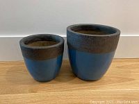 Front view of set of two blue glazed planter pots with dark brown rims sitting on wooden floor against white wall, showing size difference.