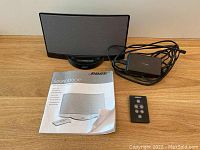 Black Bose SoundDock speaker system with remote control, power adapter, and user manual laid out on a wooden surface.