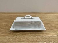 White ceramic butter dish with wood grain texture on lid, sitting on wooden surface.