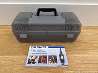 Closed grey Dremel plastic carrying case with 'Dremel' embossed on lid and a Quick Start Book laid in front.