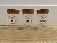 Photo showing three clear glass storage jars with cork tops arranged side by side on a wood surface. Each jar is labeled with vintage-style text: 'Things', 'Stuffs', and 'Goods'.