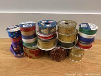 Photo showing 18 assorted rolls of ribbon and raffia in various colors, widths and patterns arranged on a flat surface.