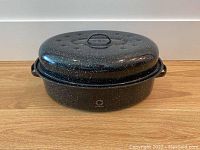 Side view of the black speckled roasting pan with its matching domed lid on top, showing the handles and lid handle.