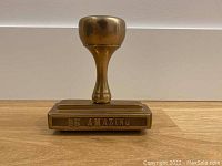Front view of the bronzed brass-effect oversized metal desk stamp featuring the phrase 'Be Amazing' engraved on the face.