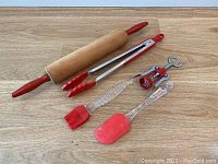 Photo of 5 kitchen utensils with red accents including rolling pin, tongs, corkscrew, silicone brush, and spatula laid out on a wooden surface.