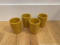 Four mustard yellow melamine tumblers placed in a semi-circle on a wooden surface against a white background. Shows the shape, size, and color of the tumblers.