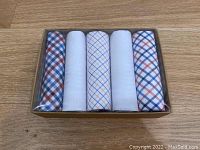 Set of 5 pocket squares with plaid and solid white patterns in box with clear lid, displayed on wood floor.