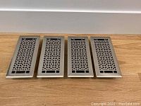 Four steel floor vent covers with cutout patterns laid out horizontally on wooden floor