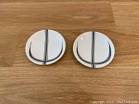 Photo of two round white trinket trays with black minimalist line designs, placed side by side on a wooden surface.