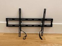 Black wall mount bracket assembled together showing horizontal mounting bracket with two vertical arms attached to it, resting against a white wall on a wooden floor.