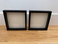 Front view of two black square shadow boxes showing the glass front and light backing, placed upright on a wooden floor.