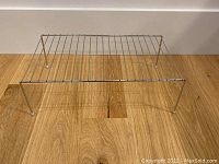 Silver metal wire rack shelf riser with four legs on a wood floor, shown from front angle.