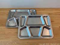 Photo showing two vintage stainless steel prison-style meal trays side by side on a wooden surface.