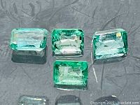 Four rectangular emerald-cut Columbian emeralds arranged in two matched pairs on a reflective dark surface, showing color and clarity.