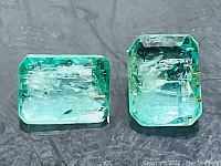 Two emerald cut emeralds side by side on a dark background showing color, clarity and size.