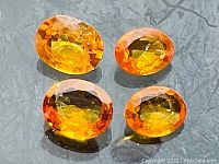 Four oval cut fire orange sapphires arranged on grey surface under reflective light showing color and cut details.