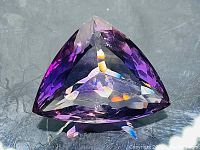 Deep purple triangular gemstone on reflective surface, oblique view showing facet reflections
