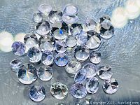 Multiple loose diamond cut Tanzanite gemstones in a cluster showing light to medium blue-purple hues on a textured surface.