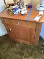Frontal view of solid wood antique cabinet showing drawer and cabinet door with white knobs and metal latch, minor surface wear and items on top.