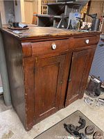 Front view of antique wooden cabinet showing two drawers with white porcelain knobs and two paneled cabinet doors below. Wood is dark stained, shows scratches and wear.