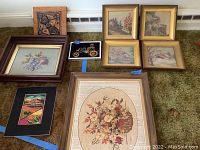 Full shot of nine vintage art and needlework pieces arranged on floor, including four gold-framed landscapes, one copper rubbing, one colorful needlework piece, and framed floral needlework.