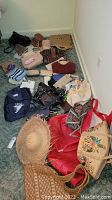 Photo shows a collection of various handbags, wallets, hats, and small cases scattered on a carpeted floor. Items include purses in pink, beige, red, and dark colours, plus white and blue striped fabric piece.