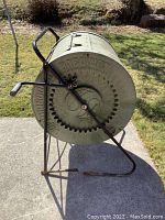 Side view of green plastic compost tumbler with black metal frame and handle showing large circular drum with embossed text and gear design.