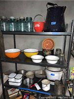 Shelf with blue glass Ball jars, red fire bucket, black speckled enamel coffee kettle on top shelf; orange and yellow enamel bowls, white enamel bowls, and some small jars on second shelf; white enamelware bucket and containers, utensils on bottom shelf.
