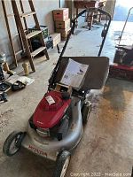 Front-side angle showing the whole Honda lawn mower with visible red engine cover, metal silver deck, front two wheels, handle bars, and a paper manual on the deck.
