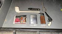 Multiple collectible lighters, clay pipe, mini toy gun and corkscrew arranged on dark surface
