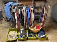 Full lot photo showing metal clothes rack with various men's clothing hanging including shirts, vests, and pants, four green plastic tubs with folded clothing and one wicker basket with folded clothing underneath the rack.